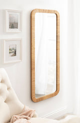 Tall rectangular wall mirror with rounded edges and a textured light wood frame, hanging on a white wall next to two small framed pictures and a cream upholstered armchair.