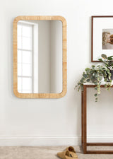 Rectangular wall mirror with rounded corners and a light wood frame reflecting a window, hung above a metal-framed console table with greenery and a framed photo.