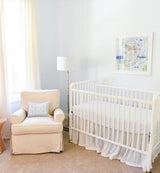 Bright nursery corner with a white crib and cushioned beige armchair featuring a monogram pillow, accented by a standing lamp and pastel Noah's Ark artwork above crib.