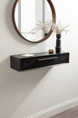Black floating console table with a single drawer mounted on a wall beneath a round mirror with a wooden frame, decorated with a black vase holding dried branches and small accessories.