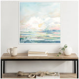 Abstract landscape painting with pastel blues, pinks, and yellows in a white frame, hanging above a wooden console table with books, a beige mug, and a white vase with greenery.