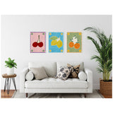 Three framed canvas artworks above a white sofa, each featuring a colorful fruit illustration with dotted border: cherries on pink, lemons on blue, and oranges on green backgrounds.