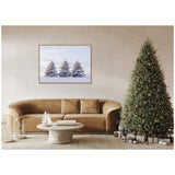 Framed canvas of three snowy pine trees against a blue sky above a brown leather sofa in a neutral-toned living room with a decorated Christmas tree and wrapped gifts.