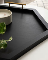 Black hexagonal decorative tray with a gold-toned handle, holding a small white candle and partially visible green plant leaves on a white surface.