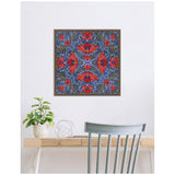 Square framed canvas print with a symmetrical kaleidoscope pattern of red poppies and green leaves on a dark blue background, hanging above a light wood desk and gray chair.