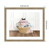 Black cat peeks over a pink towel atop a wicker laundry basket filled with folded clothes, set against a white paneled wall and wooden floor with paw prints.