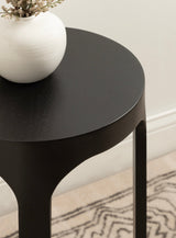 Close-up of a black, round wood drink table with smooth edges and four legs, featuring a white ceramic vase with dried flowers on top.