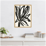 Black abstract floral brushstroke artwork by Alicia Bock in a light wood frame, hanging above a white desk with plants, books, and patterned containers.