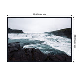 Turbulent icy blue river cascading over dark jagged rocks under an overcast gray sky, captured in a horizontal rectangular frame with black beaded trim.
