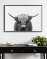 Framed black-and-white photograph of a Highland cow with long shaggy hair covering its eyes and wide horns, mounted above a black desk with books and greenery.