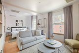 Bright modern studio with light gray velvet sofa and curtains, round nested coffee tables on textured rug, and abstract black and white art above bed.