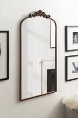Tall rectangular wall mirror with arched top and dark wood frame featuring a carved floral crest at the peak, mounted on a light wall between black-framed photos.