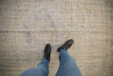 Worn vintage Oushak carpet with faded beige and gray-blue tones, viewed from above with a person wearing blue jeans and black boots standing on it.