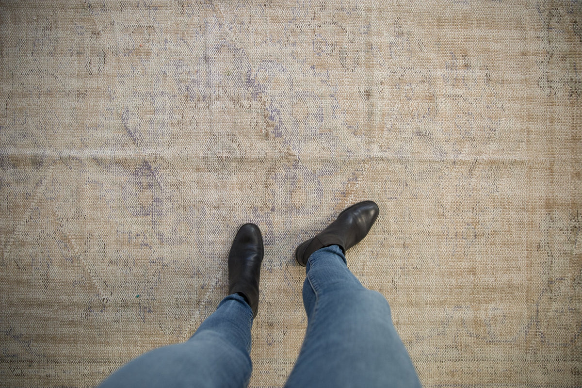 Worn vintage Oushak carpet with faded beige and gray-blue tones, viewed from above with a person wearing blue jeans and black boots standing on it.