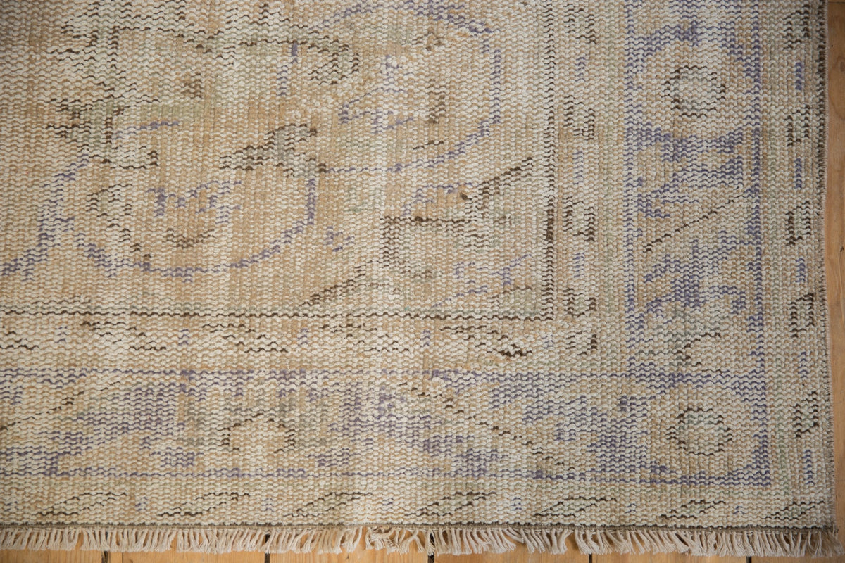 Corner of a vintage distressed Oushak carpet in beige and muted blue tones, featuring blurred geometric and floral patterns with worn texture and fringed edge.
