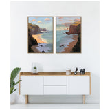 Two framed canvas paintings depicting coastal cliffs and sandy beaches under partly cloudy skies with afternoon shadows, hung side by side above a modern white wooden sideboard.