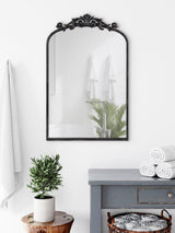 Black ornate arch mirror with floral top detail above gray distressed wooden cabinet, white rolled towels in basket, green potted plant on wooden stump in bright bathroom.