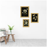 Three framed floral prints with dark backgrounds and gold frames arranged asymmetrically on a white wall above a wooden side table with a green fern in a glass vase.