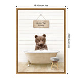 Brown bear cub sitting in a vintage clawfoot bathtub filled with bubbles against a white wooden paneled wall, with a wooden stool holding folded towels beside it.