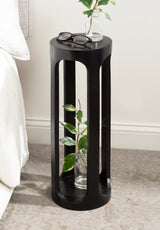 Tall, cylindrical black wood side table with elongated cutouts and a glass vase with green leaves inside, holding eyeglasses and a smaller glass vase on top