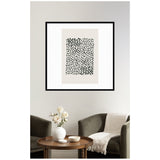 Framed abstract wall art with irregular black dots arranged in a dense rectangular cluster on a beige background, hanging above a wooden round table and two dark green armchairs.