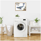 Framed canvas print of a blue whale with floral and botanical details hanging above a white front-loading washing machine in a bright laundry room.