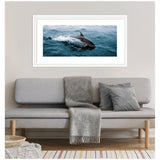 Horizontal framed photo of a dolphin skipping across blue ocean waves, displayed above a light gray sofa with a gray blanket and pillows in a minimalist living room.