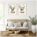 Two framed prints above a wooden bench show a front and rear view of an elephant with palm leaves and a bathtub behind it, set against a white shiplap wall.