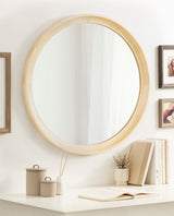 Round wall mirror with a smooth, light natural wood frame hanging above a white desk with closed and open books, small beige containers, a reed diffuser, and a small vase with dried flowers