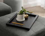 Black rectangular tray with gold handles holding a white planter with a green plant and a small gold round candle holder, resting on a gray fabric surface.