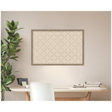 Framed beige print with a green lattice pattern hanging above a wooden desk with stacked books and a white chair, next to a tall leafy plant against a pale wall.