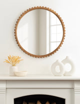 Round wall mirror with a wooden frame featuring a scalloped edge, reflecting a window; displayed above a white mantel with neutral ceramic decor and dried botanicals.