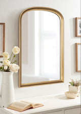 Tall arch-shaped wall mirror with a thin gold metal frame reflecting a window, hung above a white surface with an open book, flowers, and small decor.