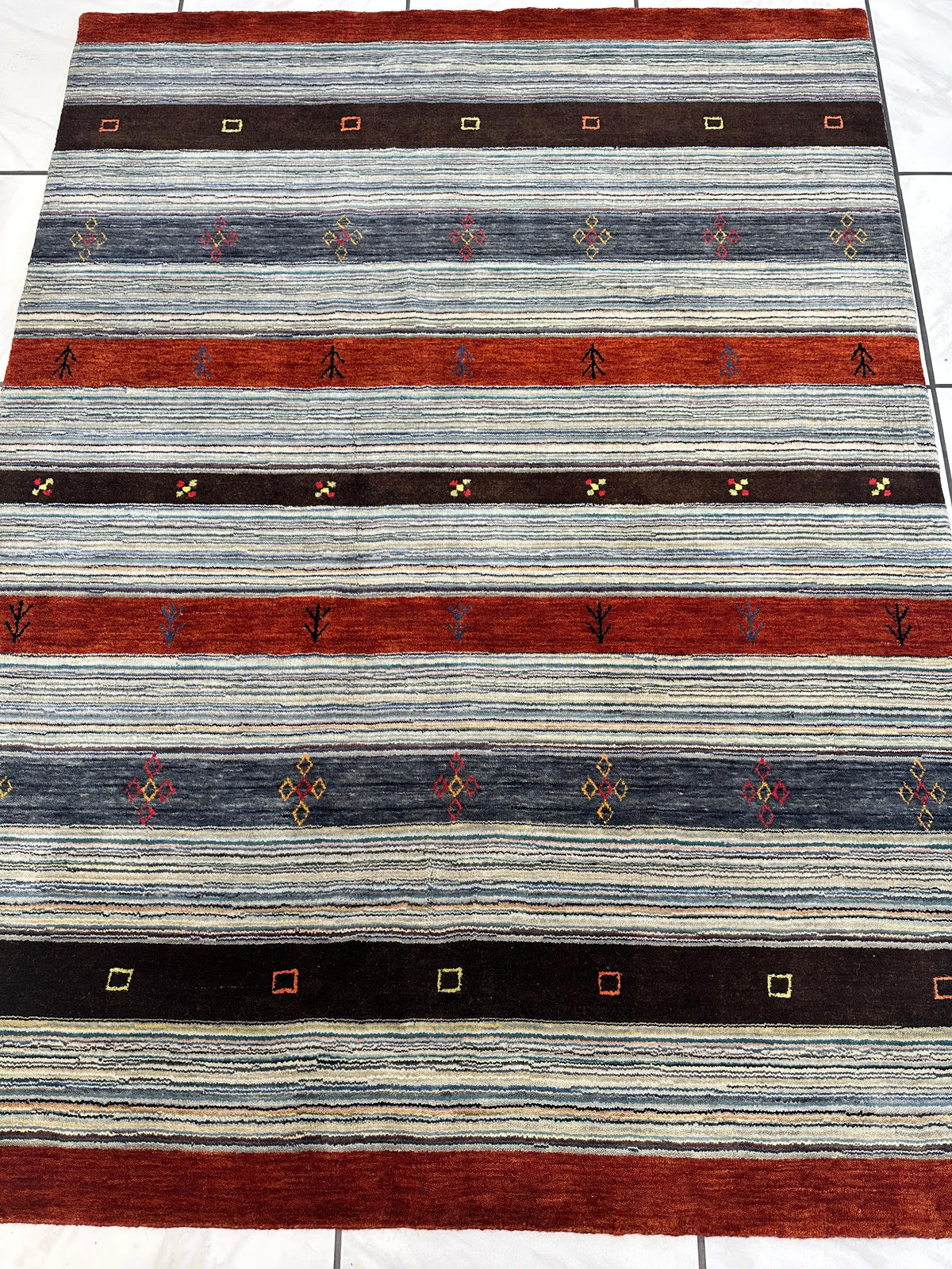 Rectangular striped rug with alternating wide bands of rust red, dark brown, and variegated gray, featuring small geometric and plant motifs in multicolored accents.