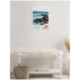 Framed canvas painting of a rocky shoreline with blue ocean waves and distant hills, hanging above a white bench with a soft fur throw and a beige bag nearby