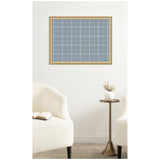 Light blue plaid fabric pinboard with white grid lines, framed in light wood, hanging on a white wall above beige upholstery chairs and a small round brass side table with a white vase.