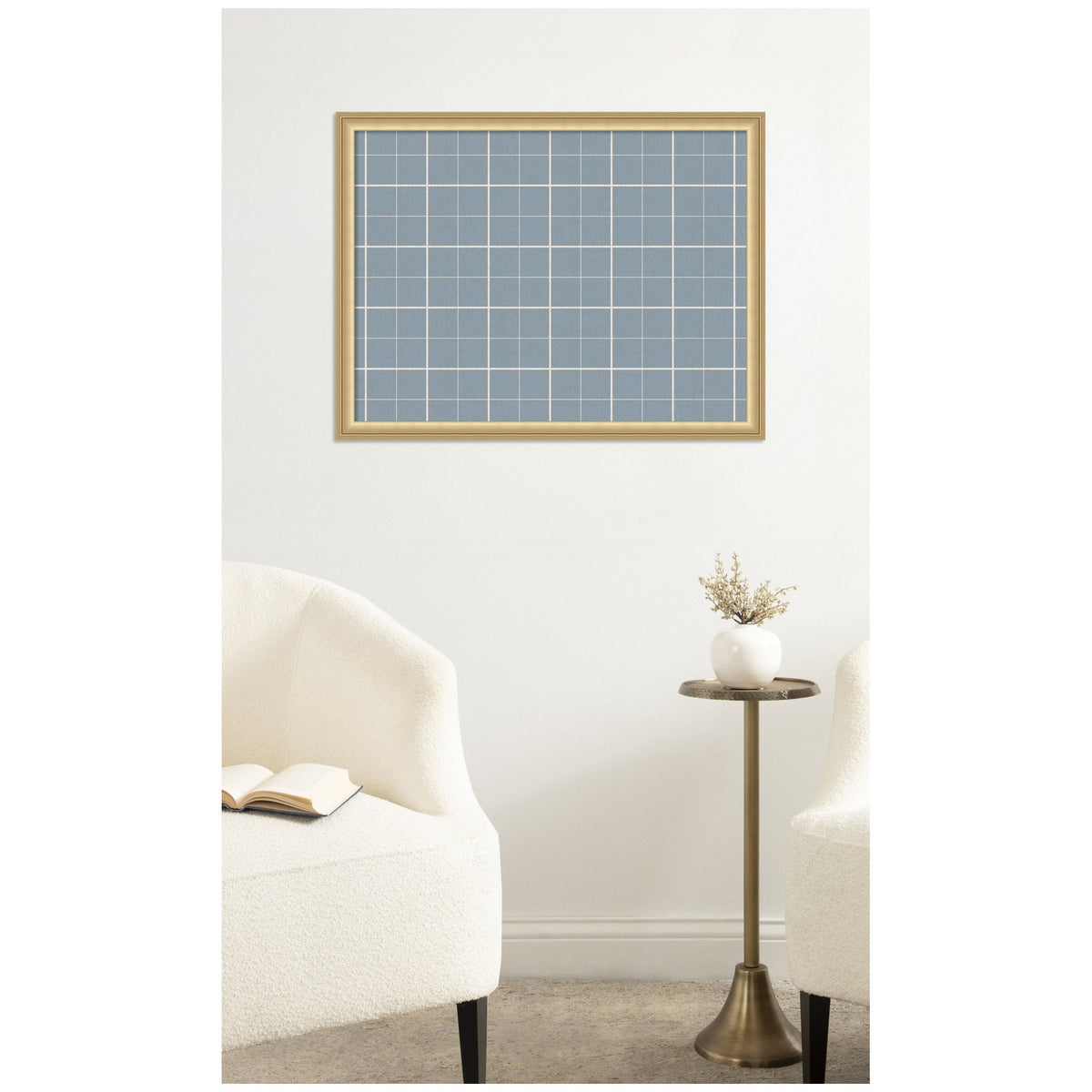 Light blue plaid fabric pinboard with white grid lines, framed in light wood, hanging on a white wall above beige upholstery chairs and a small round brass side table with a white vase.
