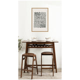 Minimalist interior scene with a dark wood bar table and two matching stools, topped with white ceramics, beneath a framed black abstract dot art on white wall.