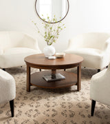 Round wood coffee table with a lower shelf holding a book, centered among four cream upholstered armchairs on a patterned beige rug with a round wall mirror above.