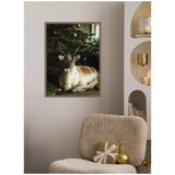 Framed canvas of a reindeer with light brown and white fur sitting under a decorated Christmas tree with gold and red ornaments, mounted on a light gray wall above a textured beige chair.