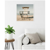 Square framed photo of a beige lifeguard tower labeled SC 3 on a sandy beach under a pale blue sky, hung on a white wall above a white sofa with light gray pillows and a wooden side table holding a green plant.