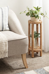 Round two-tier wicker side table with a potted green plant on top and a glass vase with greenery on the lower shelf beside a light beige upholstered sofa with wooden legs