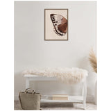 Neutral-toned watercolor painting of a brown and beige butterfly wing on textured burlap canvas, framed in wood, hanging above a white bench with a soft fur throw and gray handbag nearby.