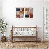 Two framed abstract geometric canvases in muted earth tones hanging above a wooden bench with white cushions, in a minimalist room with light wood floors and potted plant.