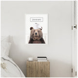 Framed canvas showing a brown bear with a toilet paper roll on its head and the text 'Just roll with it' above, hung on a white wall above a wooden bedside table with books and a cup.