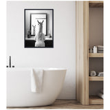 Black and white framed canvas of a llama looking into a mirror reflecting its face, hung above a white freestanding bathtub with a towel draped on the edge.