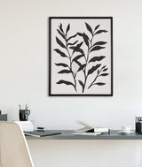 Black and white botanical silhouette print with two leafy branches in a black frame above a minimalist desk with office supplies and a white chair.