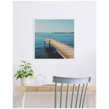 Square framed photo of a wooden pier extending into calm blue water under a clear sky, hanging above a light wood desk with a green plant and silver decorative orb.
