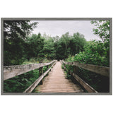Wooden bridge with rustic railings leading through dense green forest under a gray sky, framed with a simple gray border.