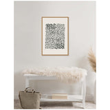 Framed abstract print with irregular black brushstroke dots arranged in a dense rectangular pattern on a white background above white bench with beige fur throw and neutral decor.